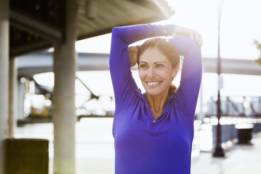 Mature woman taking a break from running to stretch her arms.  She is standing outdoors on a pedestrian walkway on the waterfront.  It is a bright, sunny day.  She is smiling, looking away from the camera.